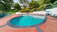 Located at 495 Laurel Hill Rd, this outdoor pool facility offers a well-appointed recreational space designed for relaxation and leisure. The centerpiece is a generously sized freeform swimming pool with crystal-clear turquoise water, accessible via multiple stainless steel entry rails and steps, making it welcoming for swimmers of all abilities.

The pool is surrounded by a broad brick paver deck finished in a warm terracotta tone, providing ample space for lounging and socializing. Numerous lightweight reclining lounge chairs are arranged along the perimeter of the deck, complemented by shaded seating areas featuring blue market umbrellas and patio table sets — ideal for guests seeking relief from the sun.

The entire area is enclosed by a combination of wrought iron fencing and low retaining walls, ensuring both safety and a sense of privacy. The backdrop features mature deciduous and evergreen trees that rise beyond the fence line, creating a natural, verdant setting that enhances the overall ambiance. The surrounding landscaping adds a lush, park-like quality to the space.

The pool area's layout is thoughtfully organized, with clear circulation paths around the pool's curved edges. The combination of open sky above, natural greenery, and the reflective quality of the water makes this an inviting and visually appealing outdoor amenity.