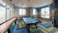 This dynamic game and social lounge is designed to energize and entertain. At its center sits a full-size pool table illuminated by a striking multi-arm pendant chandelier, while a shuffleboard table lines the adjacent wall, offering additional recreational options. The flooring makes an immediate visual statement with a bold chevron-patterned carpet in shades of blue, green, and gold, anchoring the room's lively aesthetic.

Large windows on multiple walls flood the space with natural light and offer views of the surrounding landscape, including palm trees and neighboring architecture. One corner of the room is wrapped in a richly layered typographic mural wallpaper, blending urban artistry with an eclectic, creative atmosphere. This corner transitions into a cozy lounge nook furnished with a wingback chair, ottoman, and leather sofa, perfect for relaxed conversation or unwinding.

A separate alcove features a teal accent wall with a wall-mounted flat-screen television and comfortable seating, creating an intimate viewing area within the larger space. Recessed ceiling lighting complements the pendant fixture throughout, ensuring the room is well-lit at all hours. The overall design balances bold, artistic expression with functional amenities, making it a versatile space for both social gatherings and leisure activities.