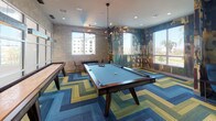 This dynamic game and social lounge is designed to energize and entertain. At its center sits a full-size pool table illuminated by a striking multi-arm pendant chandelier, while a shuffleboard table lines the adjacent wall, offering additional recreational options. The flooring makes an immediate visual statement with a bold chevron-patterned carpet in shades of blue, green, and gold, anchoring the room's lively aesthetic.

Large windows on multiple walls flood the space with natural light and offer views of the surrounding landscape, including palm trees and neighboring architecture. One corner of the room is wrapped in a richly layered typographic mural wallpaper, blending urban artistry with an eclectic, creative atmosphere. This corner transitions into a cozy lounge nook furnished with a wingback chair, ottoman, and leather sofa, perfect for relaxed conversation or unwinding.

A separate alcove features a teal accent wall with a wall-mounted flat-screen television and comfortable seating, creating an intimate viewing area within the larger space. Recessed ceiling lighting complements the pendant fixture throughout, ensuring the room is well-lit at all hours. The overall design balances bold, artistic expression with functional amenities, making it a versatile space for both social gatherings and leisure activities.