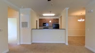 This single-floor residence offers a welcoming, open-concept layout designed for comfortable everyday living. The kitchen serves as a central focal point, featuring warm medium-toned wood cabinetry, dark granite-style countertops, and black appliances including a refrigerator, built-in microwave, and range. A peninsula counter with a decorative pendant light above elegantly separates the kitchen from the adjoining living area, creating a natural flow between spaces while maintaining a sense of openness. Structural columns flank the kitchen peninsula, adding an architectural detail that enhances the overall design. Crown molding runs throughout the space, lending a refined and polished character to the interior. The living area is finished with plush neutral carpeting that adds warmth and comfort underfoot. Walls are painted in a soft, light tone that maximizes brightness and creates an airy ambiance. A ceiling fan is visible overhead, contributing to year-round comfort. Natural and artificial lighting work together to keep the interior feeling open and inviting. The overall design balances practicality with classic detailing, making this a functional and well-appointed single-floor living space.