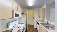 This single-floor residence offers a well-organized and efficiently designed layout, centered around a galley-style kitchen that serves as the heart of the home. The kitchen is thoughtfully appointed with light wood cabinetry lining both sides, providing generous storage space for all culinary needs. White appliances, including a gas range, over-the-range microwave, refrigerator, and dishwasher, are seamlessly integrated into the design, ensuring full functionality. A stainless steel sink is positioned along one wall, complementing the clean, neutral countertops. Overhead, a bright fluorescent light fixture ensures the space is well-lit throughout the day. Rich hardwood flooring runs the length of the kitchen, adding warmth and a sense of continuity to the space. A doorway at the end of the kitchen leads to an additional room, suggesting a connected and flowing floor plan that maximizes the use of space. The white walls and light cabinetry combine to create a bright, airy atmosphere, making the kitchen feel open and welcoming despite its compact, efficient footprint. Overall, the space is practical and move-in ready, with all the essential features needed for comfortable everyday living.