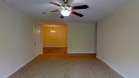This single-floor residence offers a welcoming and functional layout designed for comfortable everyday living. The living area is finished with soft, neutral-toned carpeting that adds warmth underfoot, while a ceiling fan with a built-in light fixture provides both air circulation and ambient lighting. Crown molding trims the ceiling, lending a classic architectural touch to the space. Adjacent to the living area, the dining room is distinguished by rich hardwood flooring and an elegant multi-arm chandelier that serves as a refined focal point. The transition between the two spaces is seamless, creating an open, connected feel that is ideal for both relaxing and entertaining. Warm yellow-toned walls in the dining area contrast gently with the cooler, neutral palette of the living room, adding visual interest throughout. A white paneled entry door with hardware detail provides a clean, traditional look at the entrance. White baseboards and trim throughout the space contribute to a crisp, well-maintained aesthetic. The overall ambiance is cozy yet classic, combining practical finishes with understated decorative elements to create a space that feels both livable and inviting.
