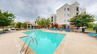 Located at 1240 Riverwalk Terr in Jenks, Oklahoma, this outdoor amenity space centers around a generously sized swimming pool with crystal-clear turquoise water. The pool area is thoughtfully designed with a spacious concrete sun deck that wraps around the pool, providing plenty of room for relaxation and recreation. Numerous blue lounge chairs and seating arrangements are distributed throughout the deck, offering comfortable spots for sunbathing and socializing. The space is enclosed by a black wrought-iron security fence, ensuring privacy and safety for guests. Mature trees, ornamental shrubs, and seasonal flowering plants are strategically placed throughout the perimeter, adding natural color and shade to the environment. The surrounding landscape opens up to open green space and scenic views beyond the fence line, including glimpses of nearby recreational facilities. The multi-story residential buildings nearby feature a classic architectural style with stucco and brick exteriors, balconies, and cornice detailing, forming an attractive backdrop to the outdoor amenity space. A dedicated clubhouse structure with a pitched roof is also visible within the complex. Overall, the pool area presents a resort-like atmosphere well-suited for outdoor leisure in a well-maintained community setting.