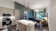 This contemporary single-floor residence is designed with a cohesive modern aesthetic, combining functionality and style throughout. The open-concept kitchen is a standout feature, boasting soaring ceilings that amplify the sense of space, light wood-tone cabinetry extending to impressive heights, stainless steel appliances, a clean white countertop, and a central island with bar seating illuminated by sleek cylindrical pendant lights. A textured dark accent wall adds visual depth, anchoring the transition between the kitchen and living areas.

The living room is bright and inviting, with floor-to-ceiling sliding glass doors that open to an outdoor patio, filling the space with natural light and views of lush greenery. Dark hardwood-style flooring runs throughout, complementing the neutral color palette.

Two well-appointed bedrooms each feature large windows with garden views, generous natural light, and ample room for comfortable furnishings. The two bathrooms are finished in a contemporary style, with wide-format tile flooring, wood-tone vanities, undermount sinks, and large framed mirrors. A dedicated in-unit laundry closet with a stacked washer and dryer adds everyday convenience to this well-designed residence.