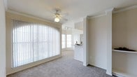 This single-floor residence offers a thoughtfully designed layout that balances openness with defined living zones. The living area is a standout space, featuring large windows dressed with vertical blinds that invite an abundance of natural light, creating a bright and airy atmosphere. Crown molding lines the ceiling, adding a refined architectural touch, while a ceiling fan with a light fixture provides both comfort and practicality. Plush carpeting runs throughout the main living areas, contributing to a warm and inviting ambiance.

A half-wall partition elegantly separates the living area from the kitchen, maintaining an open, connected feel while providing a sense of definition between the two spaces. The kitchen is equipped with track lighting and a pass-through counter, making it both functional and social. Adjacent areas include additional rooms with ceiling fans, suggesting well-ventilated and comfortable bedrooms or secondary living spaces.

The overall floor plan, as viewed from above, reveals an efficient use of space with clearly delineated rooms arranged in a cohesive, interconnected manner. The consistent neutral color palette throughout — from the white walls to the light carpeting — lends the home a clean, classic aesthetic that is both timeless and easy to personalize.