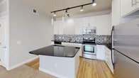 Located at 9608 E Jefferson Place in Aurora, Colorado, this single-floor residence offers a well-designed, open-concept layout that maximizes functionality and flow. The kitchen serves as a central highlight of the space, featuring white cabinetry extending to the ceiling, dark granite countertops, and a striking mosaic tile backsplash in black, white, and gray tones. Stainless steel appliances — including a range, microwave, and refrigerator — complement the modern aesthetic, while a central island with a dark countertop provides additional workspace and casual dining options. Track lighting overhead ensures excellent illumination throughout the kitchen area. Hardwood-style flooring anchors the kitchen, transitioning seamlessly into plush neutral carpeting in the open living area. The living space is generously proportioned, bathed in natural light from windows and a glass-panel exterior door that opens to the outside. The bathroom is finished with clean white tile surrounding a full soaking tub and shower combination, set on warm wood-look flooring. A spacious walk-in closet with wire shelving and hanging rods offers ample storage. Neutral gray walls and white trim throughout create a cohesive, contemporary ambiance.