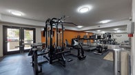 The Eagle Heights Pleasanton Fitness Center is a thoughtfully designed, single-floor gym that provides a well-rounded exercise environment. The space is anchored by a bold orange accent wall that adds energy to the room, complemented by neutral gray walls and dark rubber flooring suited for heavy equipment use. Flush-mount ceiling lights illuminate the entire area evenly throughout.

The main workout area features a prominent multi-station cable machine at its center, ideal for a wide range of strength training exercises. Alongside it, multiple treadmills and cardio machines, including an elliptical and a stationary bike, are positioned along one wall and reflected in large wall-mounted mirrors that enhance the sense of space. A dedicated free weight alcove houses a full dumbbell rack displayed against the signature orange wall, accompanied by a step platform and workout bench.

A secondary area provides additional strength equipment, including a weight bench and a cable row machine. The facility includes two restrooms, a dual-basin water fountain station with a bottle-filling option, and posted fitness center rules. A wall-mounted television and a window offering a view of the exterior greenery further enhance the experience, making this a functional and inviting place to work out.