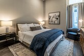 This single-floor bedroom suite is thoughtfully designed with a modern and refined aesthetic, offering a comfortable and functional living space. The bedroom is generously proportioned, featuring tall ceilings that amplify the sense of openness, and rich hardwood flooring that runs throughout, grounding the room with warmth and texture.

Natural light filters in through multiple windows dressed with floor-length grey curtains, creating a soft, welcoming ambiance. Recessed lighting and a ceiling fan with integrated lighting provide layered illumination options. A wall-mounted flat-screen TV is paired with a solid wood dresser, adding both practicality and an earthy contrast to the neutral grey-toned walls. A full-length mirror and a closet with double doors contribute to the room's strong storage and functional layout.

The en-suite bathroom continues the clean, contemporary theme with large-format light stone tile lining the walls of a full soaking tub and shower combination. A curved shower rod, built-in niche shelf, and coordinating neutral finishes create a polished, spa-inspired feel. The overall palette throughout the suite — soft whites, warm greys, and natural wood tones — is cohesive and calming, making this space both visually appealing and highly livable.