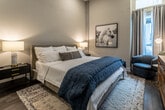 This single-floor bedroom suite is thoughtfully designed with a modern and refined aesthetic, offering a comfortable and functional living space. The bedroom is generously proportioned, featuring tall ceilings that amplify the sense of openness, and rich hardwood flooring that runs throughout, grounding the room with warmth and texture.

Natural light filters in through multiple windows dressed with floor-length grey curtains, creating a soft, welcoming ambiance. Recessed lighting and a ceiling fan with integrated lighting provide layered illumination options. A wall-mounted flat-screen TV is paired with a solid wood dresser, adding both practicality and an earthy contrast to the neutral grey-toned walls. A full-length mirror and a closet with double doors contribute to the room's strong storage and functional layout.

The en-suite bathroom continues the clean, contemporary theme with large-format light stone tile lining the walls of a full soaking tub and shower combination. A curved shower rod, built-in niche shelf, and coordinating neutral finishes create a polished, spa-inspired feel. The overall palette throughout the suite — soft whites, warm greys, and natural wood tones — is cohesive and calming, making this space both visually appealing and highly livable.
