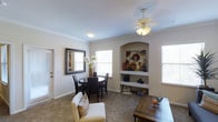 This single-story space presents a warm and inviting interior characterized by neutral tones, carpeted flooring, and classic architectural details. Crown molding lines the upper walls, adding a refined touch to the overall aesthetic. A standout feature of the main living and dining area is a built-in arched niche with open shelving, providing both a decorative focal point and functional display space. The area is illuminated by a ceiling fan with an integrated light fixture, complemented by multiple windows fitted with horizontal blinds that allow generous amounts of natural light to filter in. The dining area accommodates a round dark-wood table with matching chairs, creating an intimate setting for meals. A framed mirror on the wall enhances the sense of depth and brightness in the room. The living area flows seamlessly from the dining space, furnished with a light-colored sofa accented by patterned throw pillows. A rustic wooden coffee table adds a contrasting textural element. Overall, the space balances traditional architectural details with a comfortable, casual ambiance suited for everyday living.