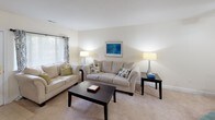 This single-floor residence offers a well-organized layout comprising two bedrooms, two bathrooms, a living room, and a kitchen. The living room is a comfortable, carpeted space featuring neutral-toned walls, plush seating arranged around a dark wood coffee table, and ambient floor lamps flanking a framed blue abstract artwork. A window with blinds allows natural light to filter in, enhancing the room's warm, inviting atmosphere.

The kitchen is equipped with white cabinetry, ample counter space, and black appliances including a refrigerator, electric range, and over-the-range microwave, creating a clean, functional workspace.

The primary bedroom features carpet flooring, windows dressed with teal ombre curtains, and a dark wood dresser with mirror. It connects to a private bathroom finished with light tile flooring, a vanity, and a linen closet. The second bedroom is also carpeted, offering a closet and a window with blinds that allows natural light in. The second bathroom features tile flooring, a shower-tub combination with a decorative curtain, and a single vanity. Throughout, the space maintains a cohesive, neutral color palette accented with teal tones.