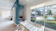 Located at 2211 Parklake Dr NE in Atlanta, GA, this single-story community clubhouse delivers a polished, contemporary amenity experience designed for both socializing and productivity. The open-concept layout is anchored by a bright, airy lounge area featuring a neutral-toned sofa, layered area rugs, and sleek accent furniture arranged beneath an elegant chandelier and shiplap ceiling. Large transom-topped glass-paned doors flood the space with natural light and provide visual connectivity between zones.

A dedicated kitchenette area offers a clean white countertop, undermount sink, under-counter refrigerator, and bar-height seating with modern wire stools — ideal for casual gatherings or catered events. A wall-mounted flat-screen TV adds to the functionality of this entertaining hub.

Adjacent to the main lounge, a game room features a full-size foosball table, a bold teal accent wall with artistic texture, and expansive floor-to-ceiling windows overlooking a landscaped outdoor courtyard with a pergola and lounge seating. A co-working nook with communal table and task seating, along with glass-enclosed private offices visible in the background, supports professional needs. Throughout, light herringbone hardwood flooring, recessed lighting, and curated artwork tie the space together with a cohesive, upscale aesthetic.