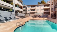 Located at 1865 N Fuller Ave in the vibrant West Hollywood neighborhood of Los Angeles, this outdoor patio and pool area serves as a welcoming communal retreat for residents. The centerpiece of the space is a generously sized, freeform swimming pool with blue tile accents and stainless steel entry rails, its crystal-clear water gleaming under the abundant Southern California sunshine.

Surrounding the pool, a broad stone-tiled deck provides ample room for lounging and socializing. A row of sleek, modern chaise lounges lines the perimeter, offering prime spots for sunbathing. The patio is further furnished with a large outdoor dining table and contemporary blue chairs, ideal for al fresco gatherings, as well as additional seating sets shaded by a striped market umbrella.

The space is framed by a multi-story residential building with private balconies, many adorned with potted plants and personal touches, adding a lush, lived-in character to the courtyard. Tiered landscaping, wrought-iron railings, and stairways connect the various levels, creating a dynamic and visually interesting backdrop. Mature trees visible above the roofline complete the setting, enhancing the sense of a private, resort-like escape in the heart of Los Angeles.