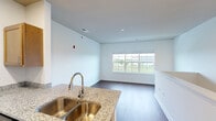This single-floor unit offers a thoughtfully designed open-concept layout that seamlessly connects the kitchen and living areas. The kitchen is a standout feature, equipped with warm honey-toned cabinetry, granite countertops, a stainless steel double-basin sink with a pull-down faucet, a full-size stainless steel refrigerator, and a built-in microwave and range. A peninsula island provides additional prep space and casual seating potential, while recessed lighting illuminates the area effectively.

The open living space flows naturally from the kitchen, highlighted by rich, dark hardwood-style flooring that runs throughout the unit. A large multi-pane window in the living area fills the space with natural light and offers views of the surrounding greenery and landscape beyond. The clean white walls and crisp trim create a bright, neutral backdrop.

The bathroom is well-appointed with a granite-topped single vanity, warm wood cabinetry, a large mirror with stylish wall-mounted lighting, and a glass-enclosed walk-in shower. The entry area features a stairwell with white balusters leading to the unit level, and a utility closet provides practical storage. Throughout, the space maintains a clean, modern aesthetic with consistent finishes and functional design.