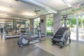 This fitness center is a comprehensively outfitted facility designed to support a full range of workout routines. The space is organized into distinct functional zones, beginning with a dedicated cardio area housing treadmills, elliptical trainers, and stationary bikes from brands such as Life Fitness. Large floor-to-ceiling windows line the perimeter, flooding the space with natural light and offering views of the surrounding greenery, creating an energizing atmosphere for workouts.

The strength and functional training zone is equally impressive, featuring a multi-station cable and weight rack system, a punching bag, medicine balls, kettlebells in a full range of weights, resistance bands, and a BOSU balance trainer. Colorful kettlebell racks and storage shelving keep equipment organized and accessible.

Mirrors are strategically placed along the walls to allow users to monitor their form throughout their sessions. The flooring throughout consists of durable, shock-absorbing rubberized material well-suited for high-impact activities. Overhead, exposed ventilation ductwork adds an industrial character to the design, while ceiling fans help maintain comfortable airflow. Bright, suspended fluorescent lighting ensures visibility across the entire facility. Double-door entryways provide easy access, and wall-mounted screens offer entertainment options for cardio sessions.