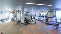 Located at 444 E. 19th Ave. in Denver, Colorado, this modern fitness center offers a well-equipped, single-floor workout facility designed to meet a wide range of exercise needs. The space features a generous open layout filled with an impressive array of cardio machines, including multiple treadmills, ellipticals, and stair climbers, all arranged in orderly rows to maximize usability and flow. Strength training equipment, including a cable machine and free weights, is also present along the perimeter, providing a comprehensive workout experience. Large windows along one wall allow abundant natural light to fill the room, creating a bright and energizing atmosphere. Recessed linear lighting in the ceiling further enhances visibility throughout the space. The vibrant, multicolored carpet adds a dynamic visual element underfoot, complementing the contemporary aesthetic of the facility. Full-length mirrors are strategically positioned to assist exercisers with form and to visually expand the space. The overall ambiance is clean, modern, and functional, making it a well-rounded fitness destination in the heart of Denver.