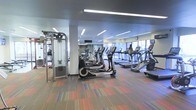 Located at 444 E. 19th Ave. in Denver, Colorado, this modern fitness center offers a well-equipped, single-floor workout facility designed to meet a wide range of exercise needs. The space features a generous open layout filled with an impressive array of cardio machines, including multiple treadmills, ellipticals, and stair climbers, all arranged in orderly rows to maximize usability and flow. Strength training equipment, including a cable machine and free weights, is also present along the perimeter, providing a comprehensive workout experience. Large windows along one wall allow abundant natural light to fill the room, creating a bright and energizing atmosphere. Recessed linear lighting in the ceiling further enhances visibility throughout the space. The vibrant, multicolored carpet adds a dynamic visual element underfoot, complementing the contemporary aesthetic of the facility. Full-length mirrors are strategically positioned to assist exercisers with form and to visually expand the space. The overall ambiance is clean, modern, and functional, making it a well-rounded fitness destination in the heart of Denver.