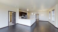 This single-floor space showcases a thoughtfully designed open-concept layout that seamlessly connects the kitchen and living areas. The kitchen is a standout feature, boasting dark espresso cabinetry that contrasts beautifully with the crisp white quartz countertops. A spacious kitchen island with an integrated sink serves as both a functional workspace and a natural gathering point, complemented by sleek pendant lighting overhead. Stainless steel appliances, including a refrigerator, microwave, and range, complete the modern culinary setup.

The rich, dark hardwood flooring flows continuously throughout the space, adding warmth and sophistication to the clean, contemporary aesthetic. Neutral white walls and smooth ceilings amplify the sense of openness, while recessed and overhead light fixtures ensure the space is well-lit throughout. A chandelier-style light fixture adds an elegant touch to the dining or living area.

Multiple doorways lead to additional rooms beyond the main living space, suggesting a well-organized floor plan with private areas for rest and convenience. The overall ambiance is modern, clean, and functional — ideal for those who appreciate a streamlined, contemporary living environment.