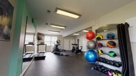 This fitness center offers a comprehensive workout environment thoughtfully organized to accommodate a wide range of exercise routines. The space is laid out across a single level with a durable rubber flooring surface throughout, providing traction and impact absorption suitable for high-intensity activities.

The cardio zone is outfitted with treadmills, an elliptical cross-trainer, and multiple spin bikes, all positioned to face a wall-mounted flat-screen television for entertainment during workouts. Natural light filters in through horizontal-blind-covered windows, complemented by recessed fluorescent ceiling fixtures that ensure consistent, even illumination across the entire space.

The strength training area features an Inspire multi-station cable machine, an adjustable weight bench, a full dumbbell rack ranging from light to heavy weights, and a TAG-branded medicine ball rack stocked with an assortment of stability and weighted balls in various sizes. Full-length mirrors line one wall, enhancing the sense of openness and allowing users to monitor their form.

Teal accent walls and green-framed mirror panels add a modern, energetic aesthetic to the otherwise neutral palette. The facility also includes restrooms and an adjacent business center accessible directly from the fitness floor, adding everyday convenience for users.