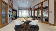 This sophisticated single-floor clubhouse lounge is designed with a contemporary flair, blending warm wood accents with a bright, airy atmosphere. The main lounge area features soaring ceilings with recessed lighting, large arched windows framed by teal drapery, and expansive glass doors that flood the space with natural light and offer picturesque views of palm trees and an outdoor pool area. Cream-toned large-format tile flooring runs throughout, unifying the open layout.

Multiple seating vignettes are thoughtfully arranged across the space. A central conversation area showcases sleek white swivel chairs grouped around a dark glass coffee table atop a circular patterned area rug. A second lounge zone features mint green upholstered chairs paired with a rich cherry wood coffee table, positioned beneath a wall-mounted flat-screen television set into an impressive floor-to-ceiling wood panel feature wall with geometric detailing.

A dedicated bar and café counter area is fitted with warm wood cabinetry, a high-gloss countertop, pendant lighting, and teal upholstered barstools. An intimate dining nook with a bold green patterned banquette and pendant lighting adds a stylish social corner. Adjacent glass-walled rooms reveal a game room with a billiards table and additional lounge seating, completing this well-appointed communal space.