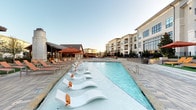 Located at 1781 W Campbell Rd in Garland, Texas, this resort-style outdoor amenity space offers a compelling blend of relaxation and entertainment. The centerpiece is a generously sized swimming pool lined with sleek in-water lounge chairs, designed for leisurely sunbathing while partially submerged. The surrounding stone-tiled deck provides ample space for additional seating and is finished with warm, neutral tones that complement the overall aesthetic.

A standout feature of the space is the covered pavilion, constructed with rich, dark wood beams and a pitched roof that lends a warm, lodge-like character. Beneath the pavilion, a floor-to-ceiling stone fireplace serves as a dramatic focal point, flanked by comfortable lounge chairs with vibrant orange cushions — ideal for cooler evenings. Adjacent seating areas include multiple outdoor dining tables with chairs, accommodating both casual gatherings and larger groups.

The pavilion also incorporates an outdoor bar area equipped with bar-height seating and a large mounted television, creating a social hub for residents. Stone masonry columns and natural flagstone flooring throughout the patio reinforce the cohesive, upscale design. Lush landscaping and wrought iron fencing frame the perimeter, providing a sense of privacy while maintaining open views of the surrounding multi-story residential buildings.