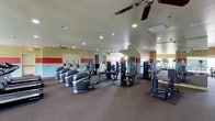 This fitness center offers a comprehensive workout environment designed to accommodate a variety of exercise needs. The space is outfitted with multiple cardio machines, including stair climbers and treadmills, alongside a selection of strength-training equipment and cable machines, providing residents with a full range of workout options. The room features a neutral carpet flooring with rubberized mats beneath the equipment for added stability and protection. Recessed lighting is installed throughout the ceiling, ensuring the space is well-lit, while multiple ceiling fans promote air circulation and comfort during workouts. The walls are painted in a combination of soft yellow and accented with bold red horizontal stripes, lending an energetic and motivating atmosphere. Large mirrors are positioned along one wall, allowing users to monitor their form. The layout is open and organized, with equipment spaced to allow for easy movement between stations. A door provides access to an adjacent area, and the overall design prioritizes functionality while maintaining a clean, welcoming environment for daily fitness routines.