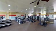 This fitness center offers a comprehensive workout environment designed to accommodate a variety of exercise needs. The space is outfitted with multiple cardio machines, including stair climbers and treadmills, alongside a selection of strength-training equipment and cable machines, providing residents with a full range of workout options. The room features a neutral carpet flooring with rubberized mats beneath the equipment for added stability and protection. Recessed lighting is installed throughout the ceiling, ensuring the space is well-lit, while multiple ceiling fans promote air circulation and comfort during workouts. The walls are painted in a combination of soft yellow and accented with bold red horizontal stripes, lending an energetic and motivating atmosphere. Large mirrors are positioned along one wall, allowing users to monitor their form. The layout is open and organized, with equipment spaced to allow for easy movement between stations. A door provides access to an adjacent area, and the overall design prioritizes functionality while maintaining a clean, welcoming environment for daily fitness routines.