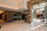 This upscale amenity floor presents a thoughtfully designed communal space that blends modern sophistication with warm, inviting finishes. The expansive main corridor and lobby area are anchored by large-format white marble tile flooring with subtle grey veining, lending a polished and refined aesthetic throughout. Warm wood-paneled walls and ceilings contrast beautifully with crisp white surfaces, creating a balanced and welcoming atmosphere.

Glass-enclosed rooms with full-height partitions provide flexible, semi-private spaces for meetings or gatherings, maintaining an open and airy feel. A dedicated lounge area features plush white sofas and a bold yellow accent chair arranged around a wall-mounted flat-screen TV, set against a dramatic dark marble feature wall with stylish globe pendant lighting overhead.

Floor-to-ceiling windows line portions of the space, allowing abundant natural light to pour in while offering views of the surrounding urban landscape. A built-in wet bar or kitchenette with open shelving, warm wood cabinetry, and a wine refrigerator serves as a central amenity hub. Additional seating nooks with round tables and mustard-upholstered chairs positioned beside large windows offer inviting spots for casual conversation. Throughout, curated artwork, lush indoor plants, and refined lighting fixtures complete the space's elevated design.