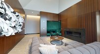 This single-floor space showcases a masterful blend of contemporary design and warm, luxurious materials. The living area immediately commands attention with its soaring vaulted ceilings, which amplify the sense of openness and airiness throughout the space. Recessed lighting is thoughtfully integrated into the ceiling, providing a soft, ambient glow that complements the natural tones of the room.

The walls are clad in rich, vertically-slatted wood paneling that runs floor to ceiling, lending warmth and a refined, organic texture to the interior. A sleek, linear built-in fireplace is seamlessly embedded within the wood paneling, serving as a striking focal point that enhances the room's sophisticated atmosphere.

A standout architectural feature is the recessed emerald green velvet banquette nook, framed by the wood paneling and accented with subtle integrated lighting. This intimate seating alcove offers a bold contrast to the neutral palette of the surrounding space, creating a vibrant yet elegant lounge area.

The main seating area features a generously proportioned, tufted modular sofa in soft grey, paired with a round, dark-framed coffee table resting atop a layered area rug. Light-toned large-format flooring ties the entire space together, reflecting light and contributing to the clean, modern aesthetic that defines this well-appointed interior.