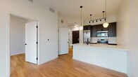 This single-floor residence showcases a contemporary design aesthetic defined by clean lines, high ceilings, and warm light hardwood flooring throughout. The open-concept layout connects the living area seamlessly to the kitchen, creating a spacious and functional living environment.

The kitchen is a standout feature, equipped with dark charcoal cabinetry contrasted by light quartz countertops and a large peninsula island with an undermount sink and matte black faucet. A geometric hex tile backsplash adds texture, while stainless steel appliances—including a range, built-in microwave, and full-size refrigerator—provide modern functionality. Pendant lighting, including globe and faceted fixtures, as well as a sleek track lighting system, illuminate the space stylishly.

The living area extends toward a glass sliding door that opens to a private balcony offering open urban views of the surrounding cityscape, filling the interior with natural light. A well-proportioned bedroom features a large window with similar exterior views and continues the hardwood flooring motif.

The bathroom maintains the unit's design continuity with a white soaking tub and shower combination, matte black fixtures, and classic basketweave tile flooring, offering a clean and timeless finish.