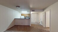 This single-floor residential unit offers a practical and functional layout designed for comfortable everyday living. The open living area is generously sized, featuring warm neutral-toned carpet underfoot and a ceiling fan with integrated lighting, creating a welcoming and airy atmosphere. The front entry opens directly into this space, reinforcing the open feel throughout.

The kitchen is efficiently designed with a galley-style configuration, featuring dark granite-look countertops, warm wood-toned cabinetry, and hardwood-style flooring. It is equipped with a white electric range, a full-size refrigerator, and a double-basin sink with ample counter space on either side. A pass-through counter connects the kitchen to the living area, enhancing the sense of openness between the two spaces.

A carpeted hallway leads to the private areas of the unit, including bedrooms and a full bathroom. The bedrooms are quiet and neutrally finished with plush carpet and clean white walls, offering a restful retreat. The bathroom is well-appointed with a dark granite-look vanity countertop, wood-toned cabinetry for storage, a large mirror with a vanity light strip, a toilet, and a bathtub. Hardwood-style flooring adds a polished touch to the bathroom. Throughout the unit, textured ceilings and central HVAC vents are consistent features, contributing to a cohesive and functional living environment.