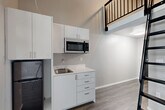 Located at 6105 Roosevelt Way NE in Seattle, WA, this two-floor loft unit offers a thoughtfully designed layout that maximizes every inch of its compact footprint. The main level opens from the entrance into a combined living, dining, and kitchen area with wood-look flooring in a cool gray tone throughout. The kitchenette is outfitted with white cabinetry, a stainless steel microwave, a single-basin sink, and a compact black refrigerator — all neatly arranged to conserve space without sacrificing functionality. A large floor-to-ceiling window floods the living area with natural light and frames views of surrounding greenery, lending an airy, open feel despite the unit's intimate scale. Soaring ceilings on the main level visually expand the space and create a dramatic vertical dimension. A sleek black metal ladder leads up to the loft on the second floor, which features its own ceiling fan and a clerestory window for additional natural light. The loft overlooks the main living area below via a black metal railing, adding an industrial-modern aesthetic. The bathroom is clean and contemporary, featuring a vessel-style sink with a dark vanity cabinet, a wall-mounted mirror, a bathtub with shower, and neutral tile flooring. This unit combines modern design with practical urban living.