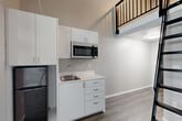 Located at 6105 Roosevelt Way NE in Seattle, WA, this two-floor loft unit offers a thoughtfully designed layout that maximizes every inch of its compact footprint. The main level opens from the entrance into a combined living, dining, and kitchen area with wood-look flooring in a cool gray tone throughout. The kitchenette is outfitted with white cabinetry, a stainless steel microwave, a single-basin sink, and a compact black refrigerator — all neatly arranged to conserve space without sacrificing functionality. A large floor-to-ceiling window floods the living area with natural light and frames views of surrounding greenery, lending an airy, open feel despite the unit's intimate scale. Soaring ceilings on the main level visually expand the space and create a dramatic vertical dimension. A sleek black metal ladder leads up to the loft on the second floor, which features its own ceiling fan and a clerestory window for additional natural light. The loft overlooks the main living area below via a black metal railing, adding an industrial-modern aesthetic. The bathroom is clean and contemporary, featuring a vessel-style sink with a dark vanity cabinet, a wall-mounted mirror, a bathtub with shower, and neutral tile flooring. This unit combines modern design with practical urban living.