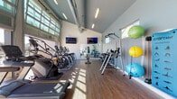 The Springs Fitness Center at 7210 Reading Rd in Rosenberg, TX is a well-appointed, single-floor facility designed to support a comprehensive workout experience. The space is defined by soaring vaulted ceilings and a dramatic wall of floor-to-ceiling glass panels that floods the interior with natural light and opens to sweeping views of the outdoor pool and surrounding community. The wood-look flooring adds warmth throughout, complementing the clean, modern aesthetic of the light gray walls.

The cardio zone lines the windowed wall, featuring a row of Matrix treadmills, ellipticals, and stair climbers positioned to take full advantage of the poolside views. Multiple wall-mounted flat-screen TVs are strategically placed throughout the space for entertainment during workouts. The strength training area is equally well-stocked, offering tiered racks of dumbbells and kettlebells in a range of weights, a weight bench, and a multi-functional cable machine. Full-length mirrors along one wall visually expand the space and aid in form correction. A colorful collection of stability and exercise balls rounds out the functional fitness options.

The entrance area includes a water fountain, a small refreshment counter with a mini refrigerator, and shelving — providing convenient amenities for visitors. Signage notes the facility is open 24 hours, making it an accessible and versatile fitness resource.