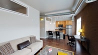 This 2-bedroom, 2-bathroom unit showcases an open-concept floor plan that efficiently combines living, dining, and kitchen areas into one cohesive space. The design draws on industrial loft aesthetics, highlighted by exposed ductwork running along the ceiling and a brick accent wall that adds texture and character to the interior.

Dark hardwood flooring runs throughout the main living area, providing a warm contrast to the light-toned walls. The living area is comfortably proportioned, offering enough space for a full sofa arrangement centered around a coffee table. A work or dining area is positioned near the kitchen, making use of available wall space with a desk setup.

The kitchen is semi-open, separated from the living area by a raised breakfast bar with seating for two to three. Warm wood cabinetry lines the kitchen walls, complemented by stainless steel appliances including a refrigerator and range. Track lighting and pendant fixtures provide targeted illumination throughout the kitchen and bar area.

Upper transom-style windows allow natural light to filter into the space, enhancing the airy, loft-like atmosphere. The overall design balances urban industrial elements with functional, everyday living features.