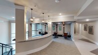 This café occupies a single level and is defined by a sophisticated, contemporary design aesthetic. At its focal point is a gracefully curved counter finished with a light marble-toned surface, serving as a natural dividing element between the entry and the main seating area. The counter's rounded form adds an architectural fluidity to the overall layout.

Overhead, a series of elegant glass pendant lights hang at varying intervals, casting warm, ambient light throughout the space. Recessed ceiling lighting complements these fixtures, ensuring consistent illumination across the room. The neutral gray walls provide a refined backdrop, accented by decorative botanical artwork and wall-mounted sconce lighting that adds a layered, welcoming glow.

The seating area is furnished with modern chairs and a cushioned bench built into the wall, creating an inviting lounge atmosphere. Plush carpeting underfoot contributes to a comfortable, quiet environment. Large windows along one wall allow natural light to filter in, enhancing the open feel of the space. A corridor visible toward the rear suggests additional areas beyond, while the overall layout balances functionality with a polished, contemporary ambiance well-suited for a café setting.
