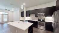 This contemporary single-floor residence showcases a thoughtfully designed open layout with a clean, modern aesthetic carried consistently throughout the space. The kitchen serves as the centerpiece, featuring rich dark espresso cabinetry that extends floor to ceiling, complemented by a textured stone tile backsplash and crisp white countertops. A center island provides additional prep space and casual seating, while pendant lighting adds a refined decorative touch overhead. Stainless steel appliances, including a range, microwave, dishwasher, and full-size refrigerator, complete the well-appointed kitchen. A built-in desk nook with matching cabinetry offers a functional workspace seamlessly integrated into the kitchen area. The living area features warm wood-style flooring, a ceiling fan with integrated lighting, and French doors that invite natural light and provide access to the outdoors. The bedroom is carpeted for comfort, with a window overlooking greenery, creating a calm and private retreat. The bathroom is finished with large-format tile flooring, a tiled tub-shower combination with decorative accent banding, dark vanity cabinetry, and a framed mirror. Recessed lighting throughout the space enhances the bright, airy atmosphere of this well-crafted home.