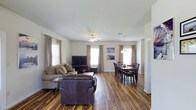 This single-floor clubhouse offers a thoughtfully designed open-concept layout that seamlessly connects the living and dining areas into one cohesive, functional space. Rich, warm-toned hardwood-style flooring runs throughout, lending an inviting character to the entire interior.

The living area is spacious and well-appointed, featuring a ceiling fan that enhances comfort and airflow. Multiple windows dressed with neutral curtains flood the room with abundant natural light, creating a bright and airy atmosphere. The layout comfortably accommodates generous seating arrangements, making it ideal for social gatherings.

The dining area flows naturally from the living space, anchored by a classic wooden dining table and chairs set beneath a stylish pendant light fixture. The continuity of flooring and the open sightlines between areas reinforce the sense of openness and ease of movement throughout the clubhouse.

White walls provide a clean, neutral backdrop that complements both the warm flooring and the furnishings, while the consistent use of natural light throughout ensures the space feels expansive and welcoming at all times. Overall, this clubhouse presents a comfortable, practical, and aesthetically cohesive environment well-suited for community gatherings and shared activities.