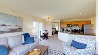 This single-floor residence offers a well-designed open-concept layout that seamlessly connects the living, dining, and kitchen areas into one cohesive and functional space. The living area is warm and inviting, featuring plush carpeted flooring, a ceiling fan with light fixture, and large sliding glass doors that bathe the room in natural light while providing direct access to an outdoor area. The kitchen is positioned along one side and features warm wood cabinetry, dark countertops, a built-in microwave, and a peninsula that doubles as a casual dining or prep space — ideal for everyday living. Adjacent to the kitchen, a dedicated dining area accommodates a table and chairs, maintaining an easy flow between cooking and entertaining. A glimpse into the bedroom reveals a comfortable and private retreat tucked just off the main living space. The overall color palette is light and neutral throughout, with cream-toned walls and carpeting that enhance the sense of brightness and openness. The thoughtfully arranged layout maximizes the use of space while maintaining a comfortable, airy ambiance suitable for day-to-day living.