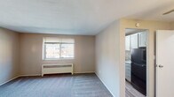 This single-floor unit offers a straightforward, functional layout designed to maximize usable space. The living area is carpeted in neutral gray tones and finished with warm beige walls, creating a clean and understated atmosphere. A window fitted with horizontal blinds allows natural light to filter in while providing an outdoor view, and a baseboard radiator beneath it ensures efficient heating. A ceiling fan centered overhead adds air circulation to the space. A hard-surface entryway transitions into the carpeted living area at the front door, with a secondary interior door providing access to additional rooms.

The kitchen is separated from the living area by a partial wall and is accessible through an open passage. It features gray cabinetry with brushed hardware, light stone-patterned countertops, a stainless steel sink, and a freestanding gas range. A black refrigerator completes the appliance package. The kitchen floor is finished in wood-look hard flooring, distinct from the carpeted living space.

The bathroom is compact and includes a tub and shower combination enclosed by a curtain, a toilet, and wood-look vinyl flooring that coordinates with the kitchen. The overall design throughout the unit is practical and cohesive, with neutral finishes that offer a versatile base for personalization.