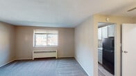 This single-floor unit offers a straightforward, functional layout designed to maximize usable space. The living area is carpeted in neutral gray tones and finished with warm beige walls, creating a clean and understated atmosphere. A window fitted with horizontal blinds allows natural light to filter in while providing an outdoor view, and a baseboard radiator beneath it ensures efficient heating. A ceiling fan centered overhead adds air circulation to the space. A hard-surface entryway transitions into the carpeted living area at the front door, with a secondary interior door providing access to additional rooms.

The kitchen is separated from the living area by a partial wall and is accessible through an open passage. It features gray cabinetry with brushed hardware, light stone-patterned countertops, a stainless steel sink, and a freestanding gas range. A black refrigerator completes the appliance package. The kitchen floor is finished in wood-look hard flooring, distinct from the carpeted living space.

The bathroom is compact and includes a tub and shower combination enclosed by a curtain, a toilet, and wood-look vinyl flooring that coordinates with the kitchen. The overall design throughout the unit is practical and cohesive, with neutral finishes that offer a versatile base for personalization.