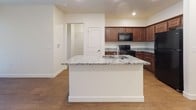 This single-floor residence showcases a thoughtfully designed open-concept layout that seamlessly connects the kitchen and living areas. The kitchen serves as a functional centerpiece, anchored by a substantial island with a striking granite countertop featuring grey and white veining, an undermount sink, and ample seating potential. Rich, warm-toned wood cabinetry lines the kitchen walls, complemented by granite countertops along the perimeter and a full suite of black appliances, including a refrigerator, range, and built-in microwave. Wood-style flooring extends consistently throughout the main living areas, adding warmth and visual continuity to the space.

The open living area is clean and bright, framed by crisp white walls and recessed lighting that provides even illumination. A keypad-secured entry door adds a modern, practical touch. The bathroom is well-appointed, featuring a wood-vanity with an integrated sink, a large mirror, built-in open shelving for storage, and a full soaking tub with a shower combination finished in white tile. A window in the bathroom allows natural light to filter in. Overall, the space presents a clean, modern aesthetic with consistent finishes throughout.