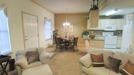 This single-story home showcases an open-concept design that integrates the living, dining, and kitchen areas into one cohesive, functional space. The living area is anchored by comfortable cream-toned seating and soft carpeting underfoot, creating a welcoming atmosphere for relaxation and entertaining.

The dining area sits centrally within the open layout, featuring a round dark-wood table surrounded by matching chairs and illuminated by an elegant chandelier overhead. Decorative greenery and subtle artwork on the warm beige walls add character to the space.

Adjacent to the dining area, the kitchen is defined by white cabinetry, a countertop peninsula that serves as a natural dividing element, and a practical sink area. The raised cabinetry above the peninsula, adorned with decorative plants, adds visual interest and vertical dimension to the space.

Natural light filters in through a window near the dining area, enhancing the overall warmth of the interior. The consistent use of neutral tones—beige walls, cream furnishings, and tan carpeting—creates a cohesive, inviting ambiance throughout this well-connected single-floor home.