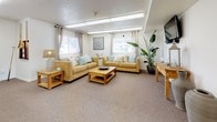 This single-floor space is thoughtfully designed to accommodate communal living and group activities across several distinct functional zones. The fully equipped kitchen features honey-toned wood cabinetry paired with light grey laminate countertops, stainless steel appliances including a refrigerator, range, microwave, and dishwasher, and a central island with additional storage and seating. Overhead fluorescent lighting ensures the workspace is well-illuminated throughout. Adjacent to the kitchen, a dedicated dining area provides seating for small groups around a round table. The spacious main gathering area serves as the heart of the space, furnished with warm golden-toned sofas accented by teal and neutral throw pillows, rustic wood coffee and side tables, and a large potted plant that adds a touch of greenery. Multiple windows dressed with sheer curtains and blinds allow natural light to filter in. A pool table occupies a prominent position in the open recreation zone, offering entertainment options. A separate, more intimate lounge nook near the entrance provides a quieter retreat with comfortable seating and decorative wall art. Neutral carpet flooring flows throughout, lending a cohesive and welcoming ambiance to the entire space.