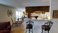 Located at 580 Pear Orchard Rd in Madison, Mississippi, this single-story residence offers a well-designed, open-concept layout that seamlessly connects the kitchen, dining, and living areas. The kitchen is equipped with dark wood cabinetry, a range with overhead ventilation, a refrigerator, and a raised breakfast bar with seating for bar-height chairs — ideal for casual meals or entertaining. Light countertops provide a clean contrast to the warm cabinetry tones. A pass-through design opens the kitchen directly to the adjoining living and dining spaces, promoting an airy, connected feel throughout. The dining area accommodates a glass-top table with ornate metal chairs, adding a decorative touch to the space. Carpeted flooring extends through the living and dining areas, contributing to a comfortable, warm ambiance. Natural light filters in through a window in the living area, brightening the open floor plan. The textured ceiling adds a classic finishing detail consistent throughout the home. Overall, this residence presents a functional and welcoming interior layout well-suited for everyday living in the Madison, MS community.