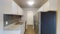 This single-floor space offers a thoughtfully designed interior that balances functionality with modern aesthetics. The galley-style kitchen is a standout feature, showcasing crisp white shaker cabinetry with brushed nickel hardware lining both sides of the space to maximize storage. Light granite countertops provide a clean, elegant work surface, complemented by a classic gray subway tile backsplash that adds a contemporary touch. Stainless steel appliances, including a full-size refrigerator, range, and dishwasher, lend a polished, cohesive look throughout the kitchen. A stainless steel sink is neatly integrated into the countertop beneath overhead cabinetry. Warm-toned wood-look flooring runs throughout, adding a welcoming feel underfoot. A flush-mount ceiling light provides consistent illumination across the kitchen. At the far end of the kitchen, a set of bi-fold doors conceals a closet or pantry, offering additional storage and keeping the space tidy. The overall layout is efficient and well-organized, making excellent use of the available space while maintaining a bright, clean ambiance throughout.