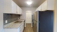 This single-floor space offers a thoughtfully designed interior that balances functionality with modern aesthetics. The galley-style kitchen is a standout feature, showcasing crisp white shaker cabinetry with brushed nickel hardware lining both sides of the space to maximize storage. Light granite countertops provide a clean, elegant work surface, complemented by a classic gray subway tile backsplash that adds a contemporary touch. Stainless steel appliances, including a full-size refrigerator, range, and dishwasher, lend a polished, cohesive look throughout the kitchen. A stainless steel sink is neatly integrated into the countertop beneath overhead cabinetry. Warm-toned wood-look flooring runs throughout, adding a welcoming feel underfoot. A flush-mount ceiling light provides consistent illumination across the kitchen. At the far end of the kitchen, a set of bi-fold doors conceals a closet or pantry, offering additional storage and keeping the space tidy. The overall layout is efficient and well-organized, making excellent use of the available space while maintaining a bright, clean ambiance throughout.