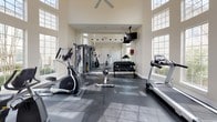 This fitness center is defined by its dramatic double-height ceilings and an expansive wall of large, grid-paned windows that flood the space with abundant natural light and offer open views of the surrounding greenery. The generous ceiling height creates an airy, open atmosphere that enhances the sense of spaciousness throughout the room.

The flooring consists of dark rubber fitness tiles, providing both durability and safety for a variety of workouts. Cardio equipment is well-represented, including treadmills, an upright stationary bike, a recumbent bike, and an elliptical trainer, all positioned to take advantage of the natural light streaming in from the oversized windows. A dedicated strength training area features a multi-station cable machine and a dumbbell rack stocked with a range of free weights. A large wall mirror spans one side of the room, visually expanding the space and supporting form-checking during exercise.

Additional amenities include a wall-mounted television, a ceiling fan, and a water cooler. Safety features such as a fire extinguisher and exit signage are clearly visible. Multiple entry and exit doors, including a glass-paned exterior door, provide convenient access. The neutral, warm-toned walls and clean layout contribute to a functional yet welcoming workout environment.