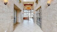 Located at 5400 Coraci Blvd in Port Orange, Florida, this sophisticated single-floor space makes a striking first impression with its grand corridor entrance clad in travertine stone walls and large-format tile flooring. The entry hall leads to an airy, open interior distinguished by soaring ceilings with richly crafted wood tray detailing and an elegant chandelier as a central focal point. A dramatic arched window above the entry doors floods the space with natural light, while wall sconces provide warm ambient illumination throughout. The main gathering area features a refined lounge with neutral-toned furnishings, a stone fireplace, and a wall-mounted display screen, creating a welcoming and functional atmosphere. A central glass-top accent table set on a marble-inlaid circular floor element serves as an eye-catching design detail. The rear of the space opens to an expansive, light-filled room enclosed by floor-to-ceiling glass window walls that frame sweeping views of a waterway lined with palm trees. This sun-drenched area accommodates both lounge and dining arrangements, offering a seamless connection to the outdoors. Throughout, the design balances coastal elegance with contemporary sophistication, creating a polished and inviting environment.