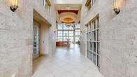 Located at 5400 Coraci Blvd in Port Orange, Florida, this sophisticated single-floor space makes a striking first impression with its grand corridor entrance clad in travertine stone walls and large-format tile flooring. The entry hall leads to an airy, open interior distinguished by soaring ceilings with richly crafted wood tray detailing and an elegant chandelier as a central focal point. A dramatic arched window above the entry doors floods the space with natural light, while wall sconces provide warm ambient illumination throughout. The main gathering area features a refined lounge with neutral-toned furnishings, a stone fireplace, and a wall-mounted display screen, creating a welcoming and functional atmosphere. A central glass-top accent table set on a marble-inlaid circular floor element serves as an eye-catching design detail. The rear of the space opens to an expansive, light-filled room enclosed by floor-to-ceiling glass window walls that frame sweeping views of a waterway lined with palm trees. This sun-drenched area accommodates both lounge and dining arrangements, offering a seamless connection to the outdoors. Throughout, the design balances coastal elegance with contemporary sophistication, creating a polished and inviting environment.